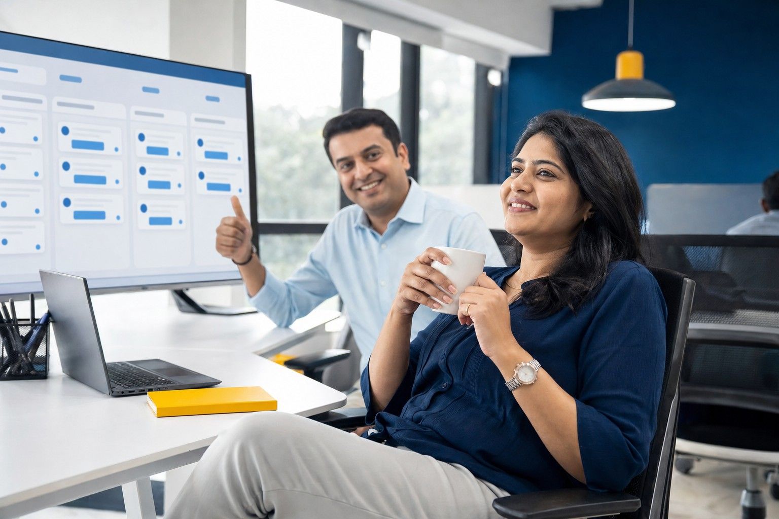 A team lead relaxing with tea while an automated workflow board runs on a large screen; a colleague gives a thumbs-up